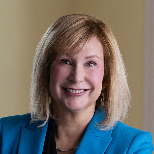Professional headshot of a smiling woman with shoulder-length blonde hair wearing a blue blazer, photographed indoors against a softly blurred neutral background.